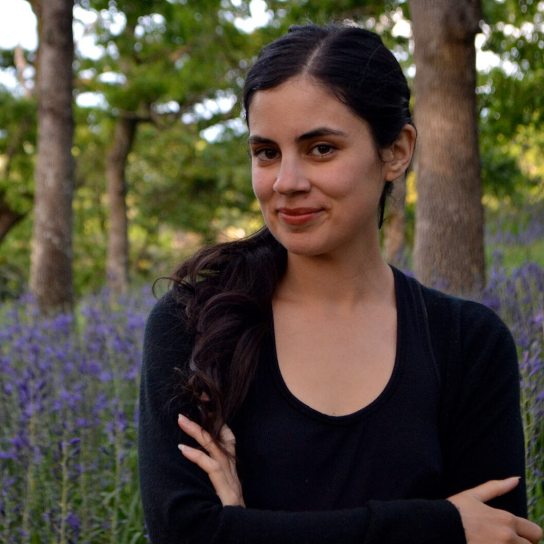Khalilah Alwani stands in front of the camas in a garry oak meadow with her arms crossed, smiling.